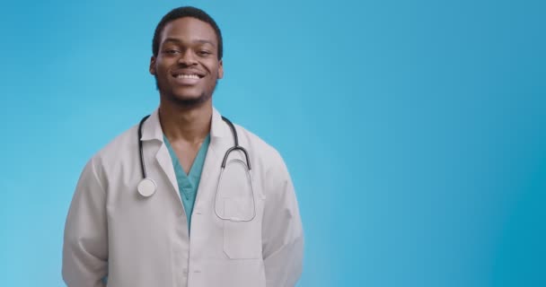 Médico americano africano confiante sorrindo e cabeça acenando, fundo de estúdio azul com espaço de cópia — Vídeo de Stock