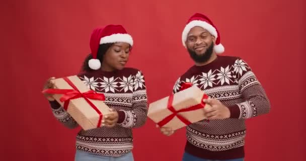 Jovem feliz casal afro-americano em Santa chapéus segurando presentes de Natal e dançando sobre fundo estúdio vermelho — Vídeo de Stock