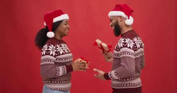 Glückliches schwarzes verliebtes Paar mit Nikolausmützen beim Austausch von Geschenkschachteln, lachend über roten Studiohintergrund — Stockvideo
