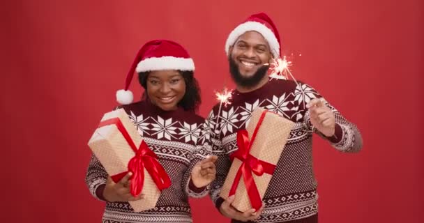 Pareja negra alegre en suéteres de invierno y sombreros de santa bailando con luces de bengala y cajas de regalo, disfrutando de la fiesta de vacaciones — Vídeos de Stock