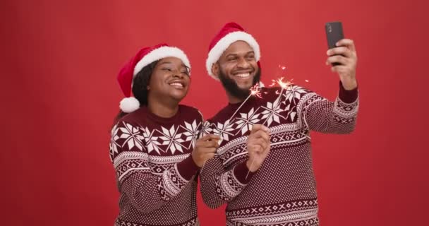 Feliz casal afro-americano em santa chapéus segurando fogos de artifício e gravação de vídeo no smartphone, fundo estúdio vermelho — Vídeo de Stock