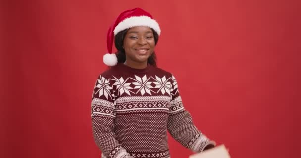 Neugierige schwarze Dame im Weihnachtsmannhut schüttelt Geschenkbox, genießt Weihnachtsurlaub, roter Hintergrund — Stockvideo