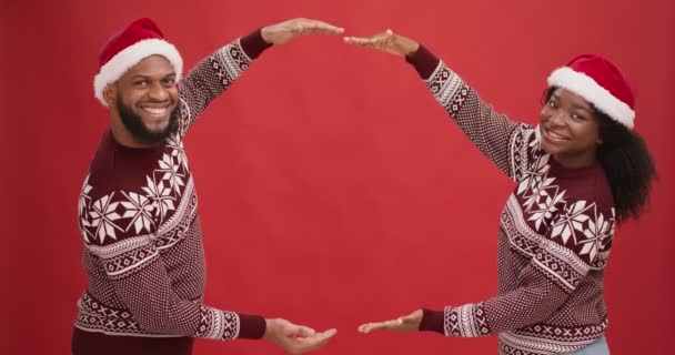 Bella offerta natalizia. Felice coppia afroamericana in cappelli di Babbo Natale rendendo cornice con le mani, sfondo rosso, spazio vuoto — Video Stock