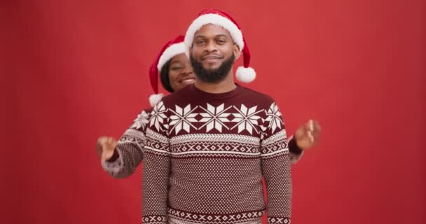 Glückliche afrikanisch-amerikanische Frau mit Weihnachtsmannhut, die Augen ihres Freundes schließt, zusammen Spaß hat, roter Studiohintergrund — Stockvideo
