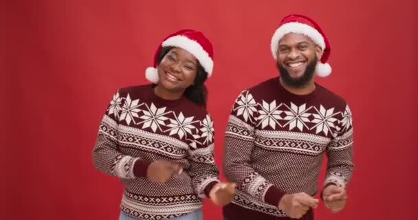 Jovem casal afro-americano vestindo camisolas e chapéus de Papai Noel dançando e estalando os dedos juntos, fundo vermelho — Vídeo de Stock