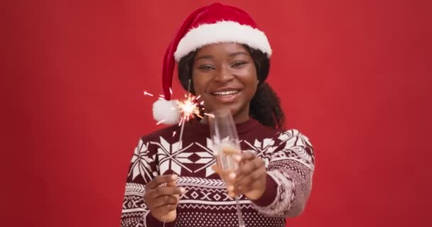 Fröhliche afrikanisch-amerikanische Dame mit Weihnachtsmütze jubelt in die Kamera mit Bengallicht und Champagnerglas, Frohe Weihnachten — Stockvideo