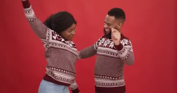 คู่รักชาวแอฟริกัน อเมริกัน ใส่เสื้อกันหนาวปีใหม่ เต้นรําอย่างมีพลัง สนุกกับปาร์ตี้คริสต์มาส พื้นหลังสีแดง — วีดีโอสต็อก