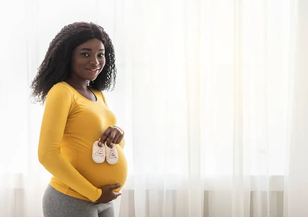 Heureuse africaine attendant femme montrant de petites chaussures pour bébé à venir — Photo