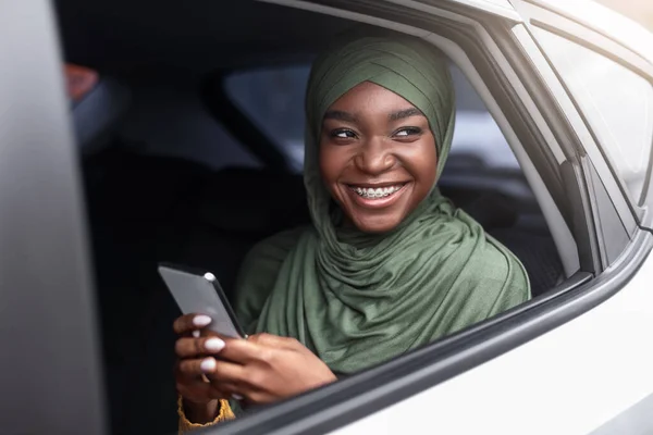 Positieve Afrikaanse moslim dame met behulp van smartphone tijdens het hebben van een road trip in de auto — Stockfoto