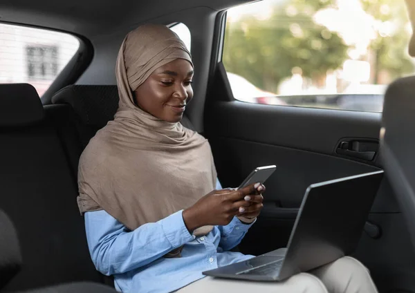 Zwarte islamitische zakenvrouw met behulp van smartphone en laptop, gaan naar kantoor met de auto — Stockfoto