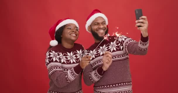 Glückliches afrikanisch-amerikanisches Paar in Weihnachtsmannmützen mit Feuerwerk und Videoaufzeichnung auf Smartphone, roter Studiohintergrund