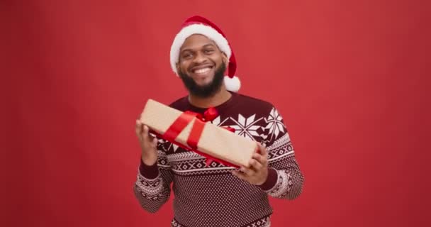 Szczęśliwy Afroamerykanin facet w zimowym swetrze i kapelusz Santa taniec z pudełka świąteczne, próbuje odgadnąć prezent — Wideo stockowe