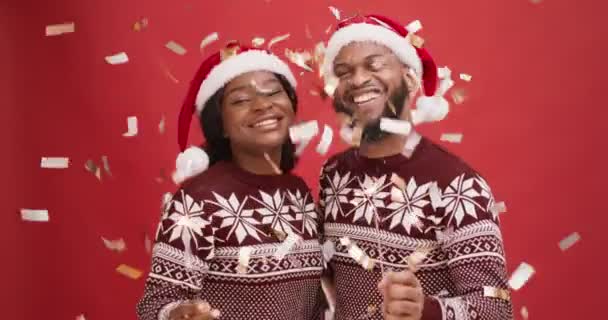 Jeune couple afro-américain célébrant Noël, dansant avec des lumières bengales unter confettis dorés, fond rouge — Video