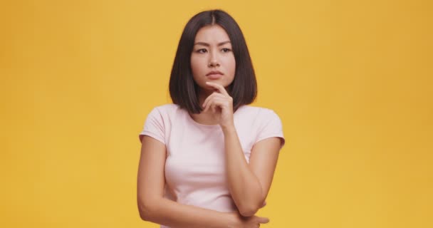 Young thoughtful asian woman pondering about something, touching her chin, orange studio background — ストック動画