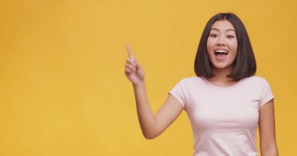 Jonge aziatische dame met idee, wijzende vinger omhoog, staande over oranje studio achtergrond met lege ruimte — Stockvideo