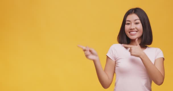 Heureuse excitée femme asiatique montrant espace vide, pointant de côté avec les deux mains, fond studio orange — Video