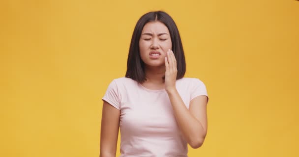 Junge asiatische Dame fühlt plötzliche Schmerzen, berührt ihre Wange und runzelt die Stirn, orangefarbener Studiohintergrund — Stockvideo