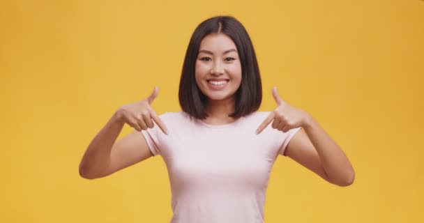 微笑着自信的年轻亚洲女人，指指点点自己，看着白色T恤上空旷的空间 — 图库视频影像