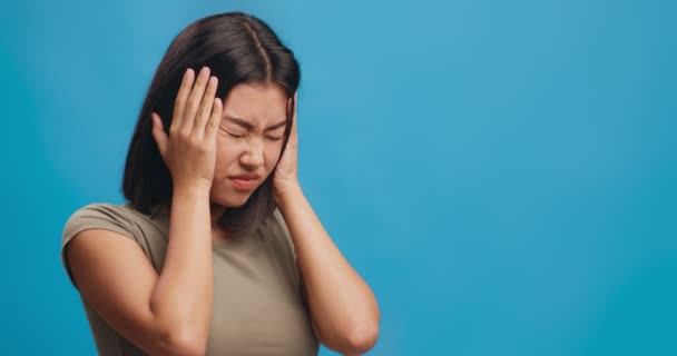 Junge Asiatin mit Kopfschmerzen, die unter Lärm leidet, Ohren mit Handflächen schließt, blauer Hintergrund — Stockvideo