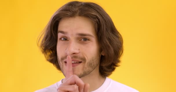 Playful young man showing hush gesture, putting finger on lips and pointing to camera, orange studio background — Stock Video