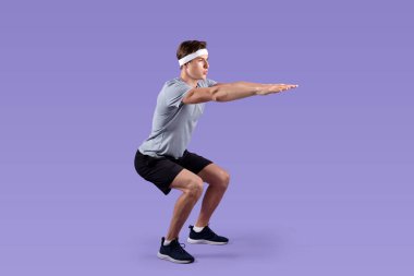 Spor giyimli atletik genç adam leylak rengi stüdyo geçmişi üzerine çömelme egzersizleri yapıyor.