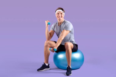 Spor elbiseli, spor elbiseli, spor salonunda oturan ve Violet stüdyosunun arka planında dambıllarla çalışan mutlu milenyum çocuğu.