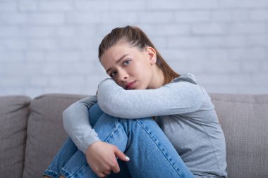 Depresyonda olan genç bir kadın evdeki kanepede dizlerine sarılıyor, olumsuz düşüncelere dalmış, akli dengesi bozuk.