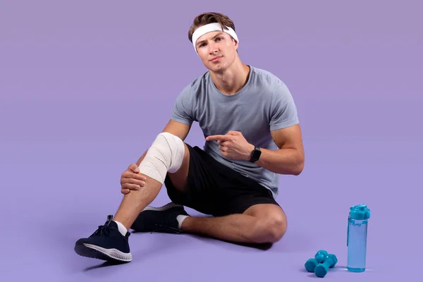 Pria jutawan atletik mengalami cedera olahraga, menunjuk pada lututnya terkilir perban atas latar belakang studio ungu — Stok Foto