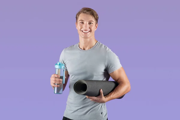 Bello atleta millenario in possesso di tappetino yoga e bottiglia con acqua su sfondo viola studio — Foto Stock