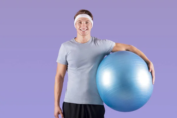 Alegre atleta milenario con pelota de fitness sonriendo y mirando a la cámara en el fondo del estudio lila — Foto de Stock