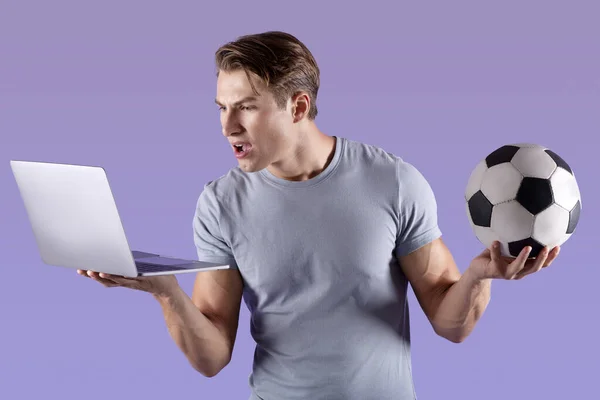 Sportieve duizendjarige man teleurgesteld in zijn voetbalteam nederlaag, kijken naar voetbalwedstrijd op laptop, violette studio achtergrond — Stockfoto