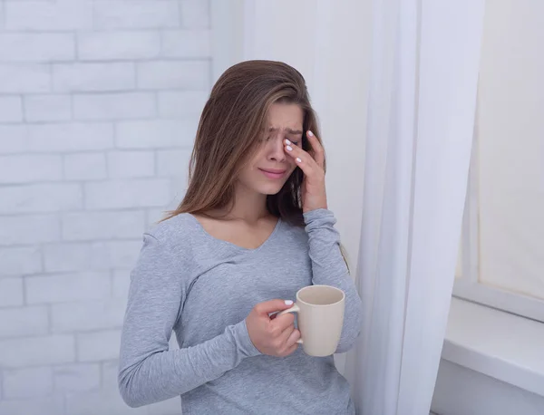 Mujer joven triste con taza de café llorando cerca de la ventana, limpiando sus lágrimas en casa — Foto de Stock