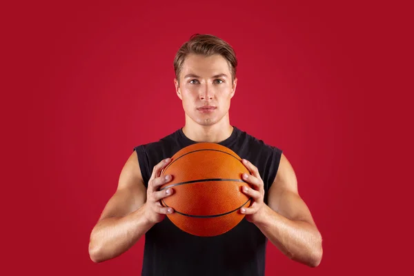 Portrait of determined young basketballer holding ball and looking at camera on red studio background — Stok Foto