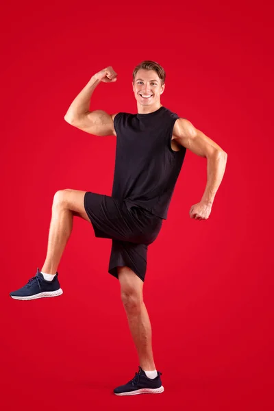 Full length portrait of young sportsman marching or walking with his knees up on red studio background — Stock Photo, Image