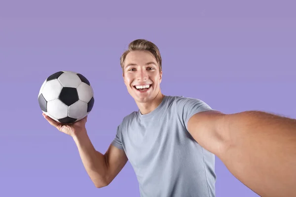 Sonriente joven con pelota de fútbol tomando selfie en su teléfono móvil sobre fondo de estudio violeta — Foto de Stock