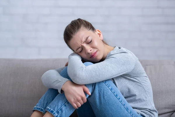 Joven mujer deprimida sentada en el sofá con los ojos cerrados y abrazando sus rodillas, sintiéndose desesperada y estresada — Foto de Stock