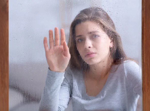 Depressed young lady touching window glass with rain drops, feeling unhappy or melancholic