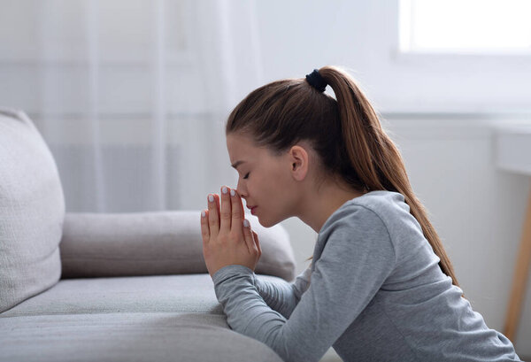 Depressed young woman praying to God, asking for help or consolation near sofa indoors, empty space