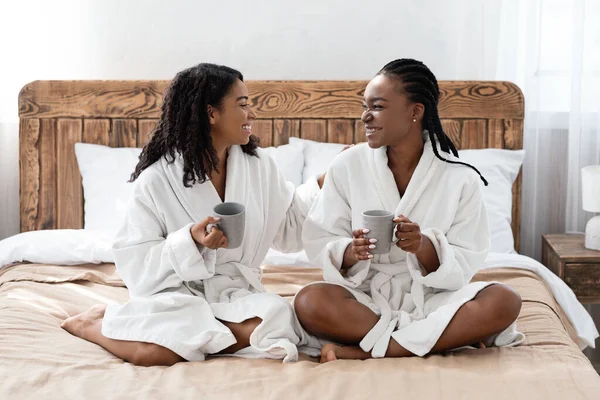 Positieve zwarte vriendinnen in badjassen zitten op bed, praten — Stockfoto