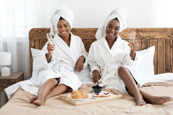 Joyful namoradas negras em roupões de banho bebendo champanhe no quarto — Fotografia de Stock