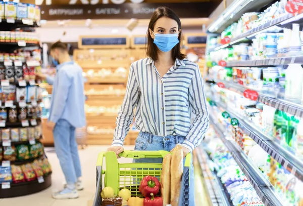 Jonge vrouw in het gezicht masker winkelen in supermarkt met kar — Stockfoto