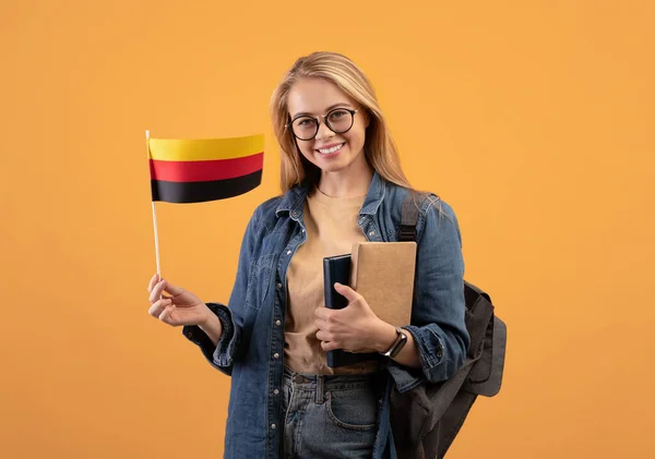 Jovem estudante loira feliz em casual com óculos, mochila e livros segurando pequena bandeira da Alemanha — Fotografia de Stock