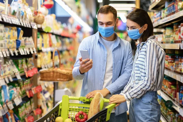 Young couple in masks with phone shopping in supermarket — Stock Photo, Image