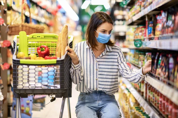 Νεαρή γυναίκα με ιατρική μάσκα με καλάθι ψώνια στην αγορά — Φωτογραφία Αρχείου
