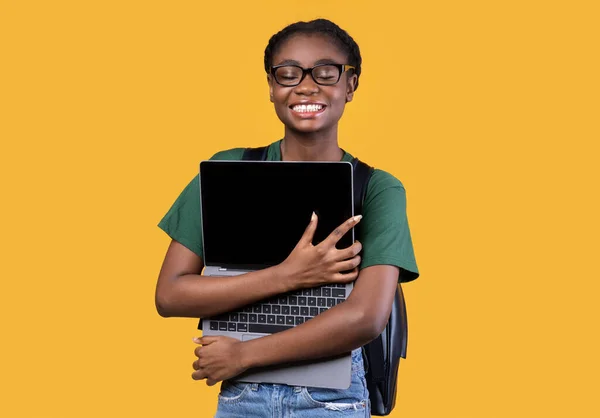 Feliz negro estudiante femenino abrazando portátil de pie sobre fondo amarillo — Foto de Stock