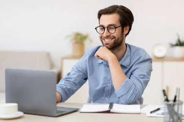 Dizüstü bilgisayarda çalışan gülümseyen iş adamı ofisinde oturuyor.