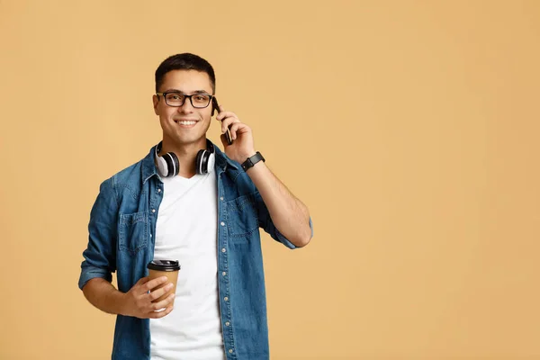 Ocupado sorrindo jovem estudante homem em óculos com fones de ouvido segurar copo de papel com café — Fotografia de Stock