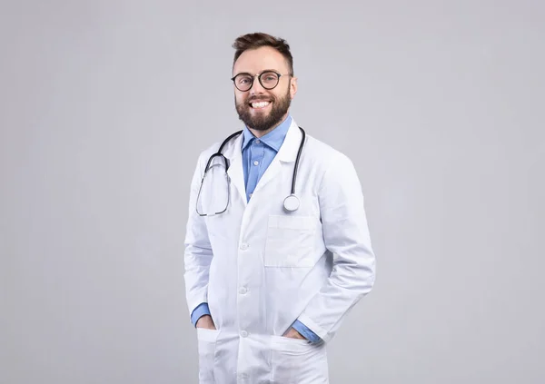Confiado médico varón caucásico en uniforme blanco sonriendo y posando sobre fondo gris estudio — Foto de Stock