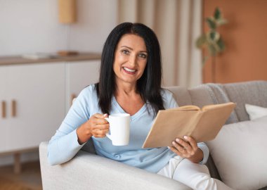 Elinde bir fincan kahve ve kitap tutan olgun bir kadın.