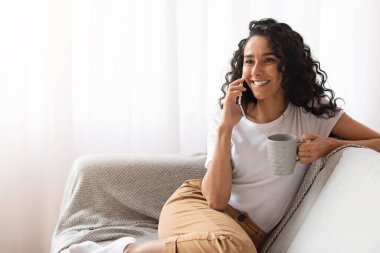 Ev kıyafetleri içinde neşeli bir bayan telefonda konuşuyor, çay içiyor.